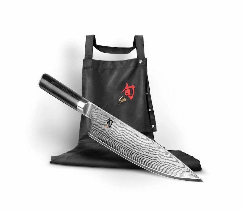 Ein KAI Weihnachtsangebot 2-teiliges Geschenkset - Shun Classic - limitiertes 1.200 Stk. Set, bestehend aus einem Shun Classic Kochmesser mit aufwendigem Damaszenerstahlmuster und einer schwarzen Canvas-Schürze mit rotem Shun-Logo auf der Brust, wird vor einem schlichten weißen Hintergrund präsentiert. Das Messer hat einen eleganten schwarzen Griff, der die Exklusivität dieses Sets in limitierter Auflage unterstreicht.