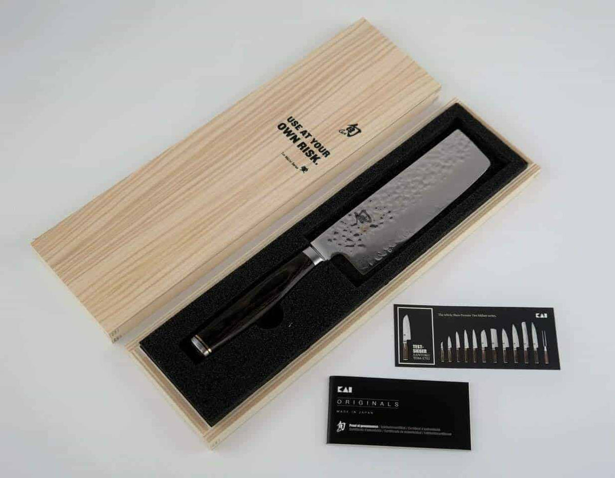Ein hochwertiges Kochmesser, das Kai Shun Premier Tim Mälzer Nakiri Messer, TDM-1742, mit einer 14 cm langen Klinge aus 32 Lagen Damaszenerstahl, wird in einer offenen Holzbox mit schwarzer Schaumstoffeinlage präsentiert. Das japanische Küchenmesser hat eine strukturierte Klinge aus rostfreiem Stahl und einen dunklen Griff. Neben der Box liegen zwei Informationskarten mit Angaben zur Marke des Messers.