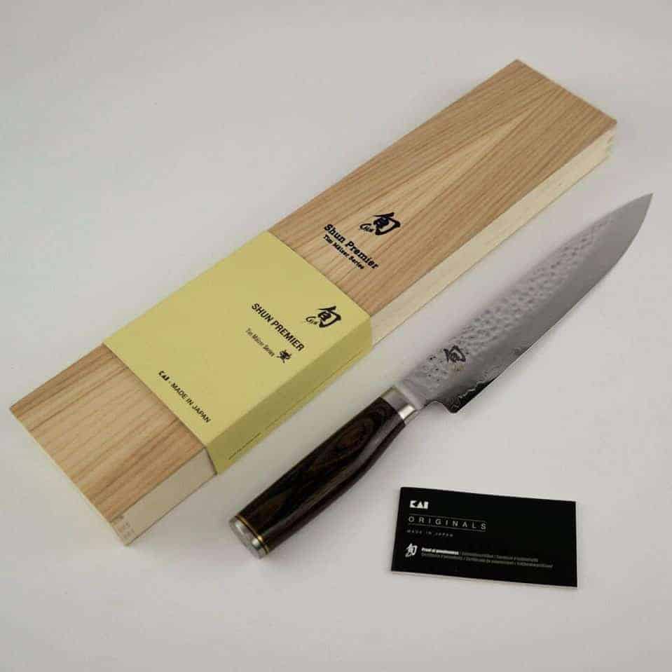 Ein hochwertiges Kai Shun Premier Tim Mälzer TDM-1704 ultrascharfes japanisches Schinkenmesser mit strukturierter Damaststahlklinge und dunklem Holzgriff wird neben seiner Holzbox-Verpackung ausgestellt. Die Box zeigt japanische Schriftzeichen und Markenzeichen sowie eine Visitenkarte mit der Aufschrift „KAI Originals“. Zusätzlich ist ein massives 30x18 cm großes Schneidebrett aus Fassholz enthalten.