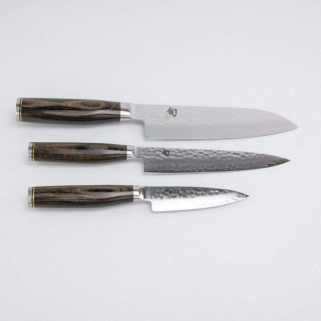 Drei handgefertigte Tim Mälzer Profimesser aus der Kai Shun Premier Kollektion mit dunklen Holzgriffen liegen horizontal auf hellem Untergrund. Die Klingen weisen unterschiedliche Muster auf: Das obere Messer hat eine glatte Klinge, das mittlere Messer hat eine gehämmerte Oberfläche und das untere Messer hat eine strukturierte Oberfläche.