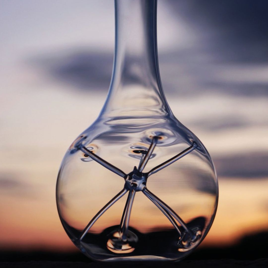 Eine Nahaufnahme des Weindekanters "Star" - Exklusive Karaffe mit Sternendesign (34 cm, Zieher), dessen klarer Glassockel mit einem Sternmotiv vor einem verschwommenen Sonnenuntergangshimmel im Hintergrund funkelt.
