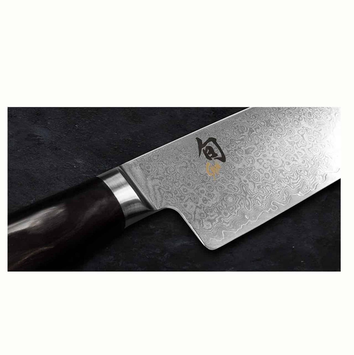 Nahaufnahme eines hochwertigen Küchenmessers mit dunklem Griff und einer glänzenden, strukturierten Damaststahlklinge mit komplizierten Mustern. Die Klinge, ein Kai Shun Premier Minamo TMM-0700 Officemesser 9 cm, weist am unteren Ende japanische Schriftzeichen auf, die auf die Marke und die Handwerkskunst hinweisen. Der Hintergrund ist eine dunkle, strukturierte Oberfläche.