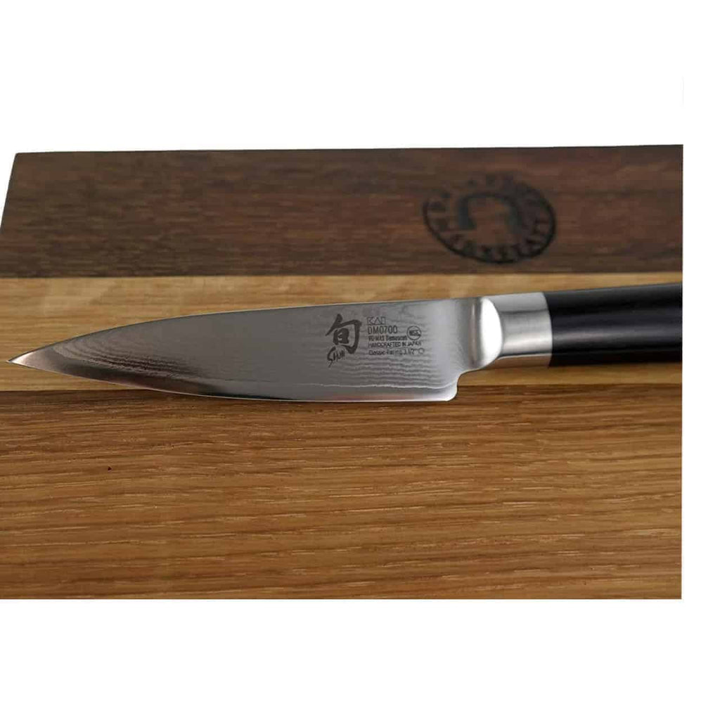 Ein Kai Shun Classic DM-0700 Officemesser mit schwarzem Griff liegt auf einem aus Weinfassholz gefertigten Schneidebrett. Die 9 cm lange Klinge aus Damaststahl ist mit eingravierten Texten und Symbolen versehen, die Marke und Typ angeben. Der Hintergrund ist schlicht und weiß.