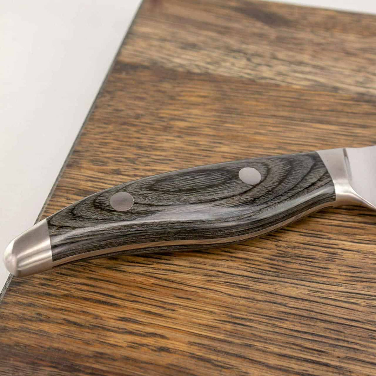 Eine Nahaufnahme des Griffs eines handgeschmiedeten Santoku-Messers KAI Shun Nagare NDC-0702, der auf einem basaltgrauen Schneidebrett aus Eiche ruht, 34 x 21 cm. Der polierte Holzgriff mit seiner glatten Oberfläche und zwei Metallnieten ist deutlich zu sehen, zusammen mit einer Teilansicht der Klinge und des Kropfes. Die Holzmaserung und die Textur des Griffs sind deutlich sichtbar.