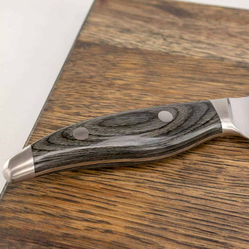 Eine Nahaufnahme des Griffs eines handgeschmiedeten Santoku-Messers KAI Shun Nagare NDC-0702, der auf einem basaltgrauen Schneidebrett aus Eiche ruht, 34 x 21 cm. Der polierte Holzgriff mit seiner glatten Oberfläche und zwei Metallnieten ist deutlich zu sehen, zusammen mit einer Teilansicht der Klinge und des Kropfes. Die Holzmaserung und die Textur des Griffs sind deutlich sichtbar.