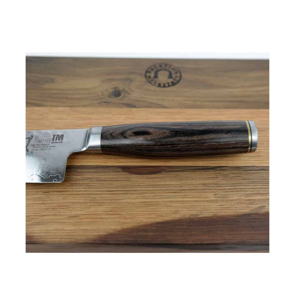 Nahaufnahme eines Kai Shun Premier Tim Mälzer TDM-1723 Messers mit detailreichem, dunklem Holzgriff, das auf dem 30x18 cm großen Küchenschneidebrett von Fassholz ruht. Die Klinge aus 32-lagigem Damaststahl weist aufwendige Gravuren in Griffnähe auf. Das Schneidebrett ist mit einem kreisförmigen Logo versehen.