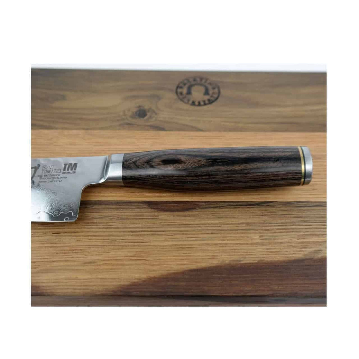 Nahaufnahme eines Kai Shun Premier Tim Mälzer TDM-1723 Messers mit detailreichem, dunklem Holzgriff, das auf dem 30x18 cm großen Küchenschneidebrett von Fassholz ruht. Die Klinge aus 32-lagigem Damaststahl weist aufwendige Gravuren in Griffnähe auf. Das Schneidebrett ist mit einem kreisförmigen Logo versehen.