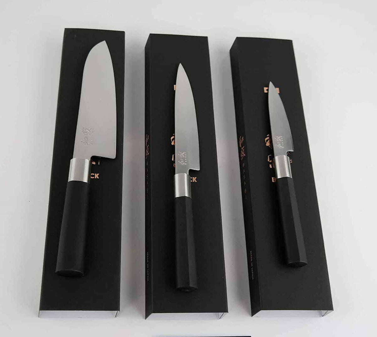 Ein Set mit drei Messern aus der Kai Wasabi Black-Kollektion, bestehend aus einem Santokumesser (16,5 cm), einem Allzweckmesser (15 cm) und einem Officemesser (10 cm), wird in eleganten schwarzen Displayboxen präsentiert. Jedes Messer verfügt über eine silberne Klinge mit schwarzem Griff, ergänzt durch einen polierten Metallkropf, der die Klinge nahtlos mit dem Griff verbindet. Zu diesem eleganten Set gehört auch ein Schneidebrett mit den Maßen 25 x 15 cm, das den raffinierten Stil von Kai Wasabi Black verk