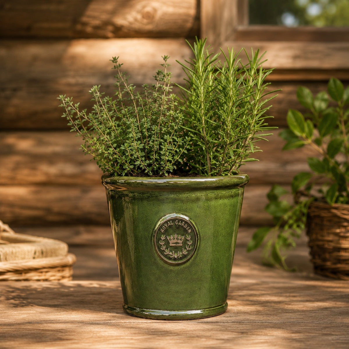 Der glasierte Pflanzkübel „Garden Green“ (Höhe 19 bis 38 cm) mit Kräutern wie Rosmarin und Thymian steht auf einer Holzfläche im Freien; das Sonnenlicht wirft Schatten auf die rustikale Holzwand im Hintergrund.