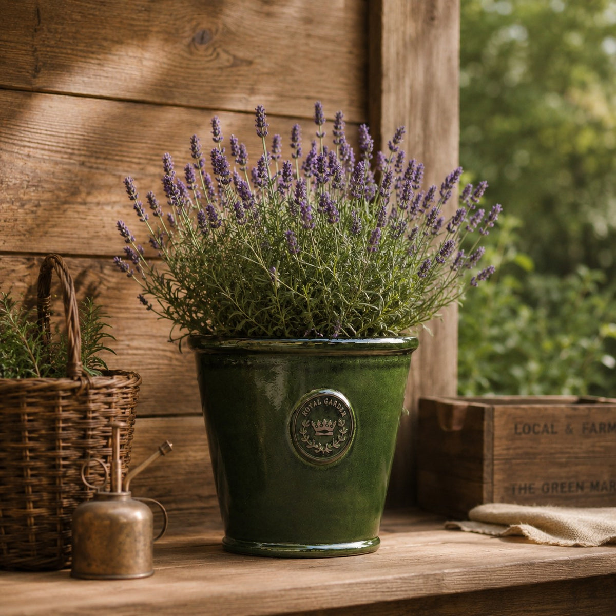 Der glasierte Pflanzkübel „Garden Green“ (Höhe 19 bis 38 cm) mit blühendem Lavendel steht auf einem Holzregal neben einem Weidenkorb, einer kleinen Messing-Gießkanne und rustikalen Kisten in einer bezaubernden Gartenkulisse.