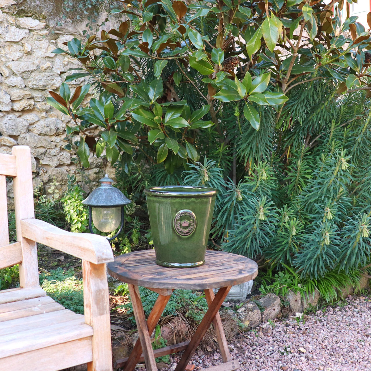Der glasierte Pflanzkübel „Garden Green“ (Höhe 19 bis 38 cm) steht auf einem kleinen runden Holztisch neben einer Holzbank im Garten, in dessen Nähe sich üppige Pflanzen und eine Steinmauer befinden. Hinter dem Tisch hängt eine Außenlaterne.