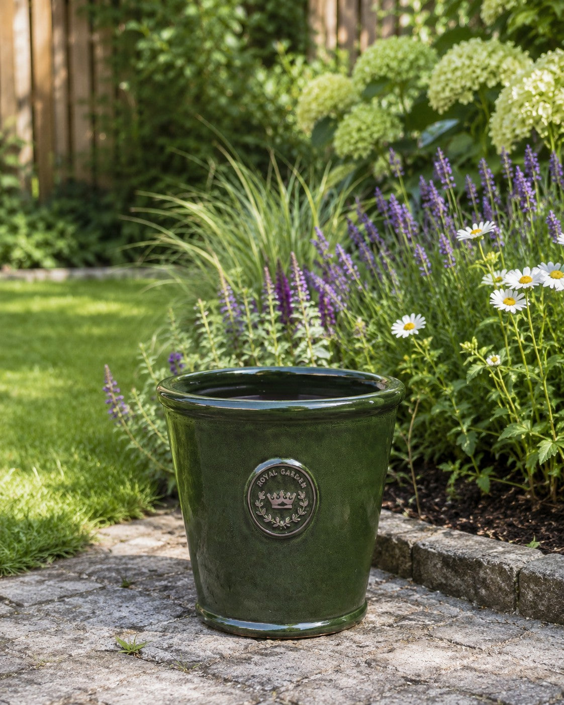 Der glasierte Pflanzkübel „Garden Green“ (Höhe 19 bis 38 cm) steht glänzend und dunkel auf einer Steinterrasse, umgeben von blühenden Gänseblümchen, violetten Blumen, üppigem Grün, Hortensiensträuchern und einem Holzzaun – eine idyllische Gartenszene.