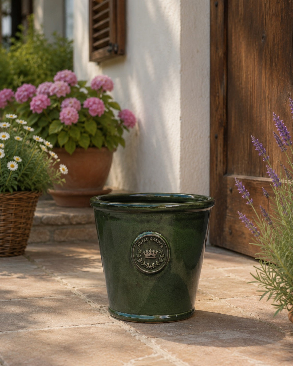 Der glasierte Pflanzkübel „Garden Green“ (Höhe 19–38 cm) steht auf einer Steinterrasse in der Nähe einer Holztür, umgeben von blühenden Blumen in Terrakottatöpfen und geflochtenen Körben.