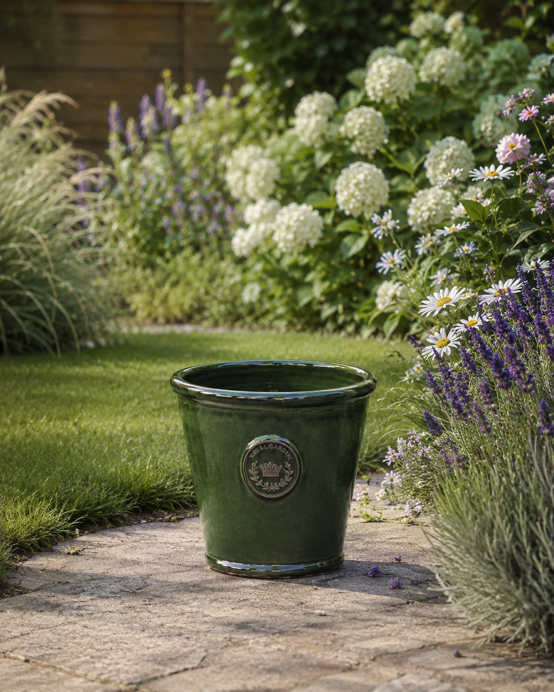 Der glasierte Pflanzkübel „Garden Green“ (Höhe 19 bis 38 cm) steht auf einem Steinweg und ist umgeben von weiß blühenden Hortensien, violetten Blumen, Margeriten sowie grünem Gras im Hintergrund.