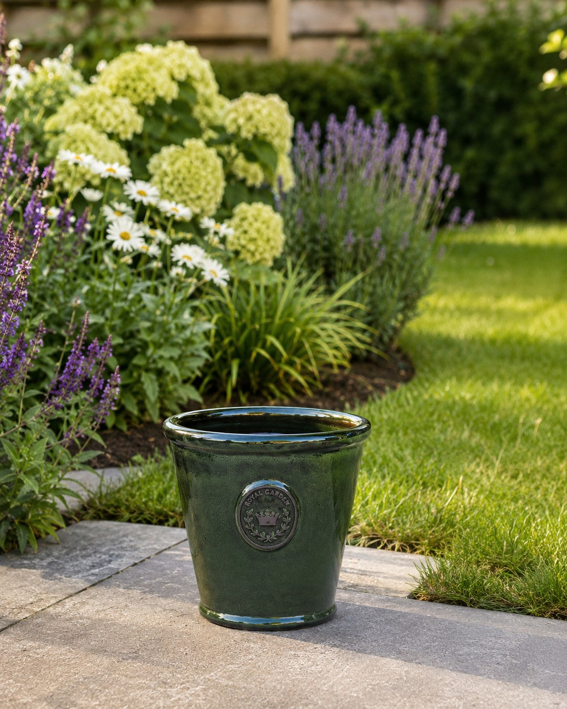 Der glasierte Pflanzkübel „Garden Green“ (Höhe 19 bis 38 cm) steht auf einer Steinterrasse im Garten, umgeben von blühenden Hortensien, Margeriten, Lavendel und saftig grünem Gras.