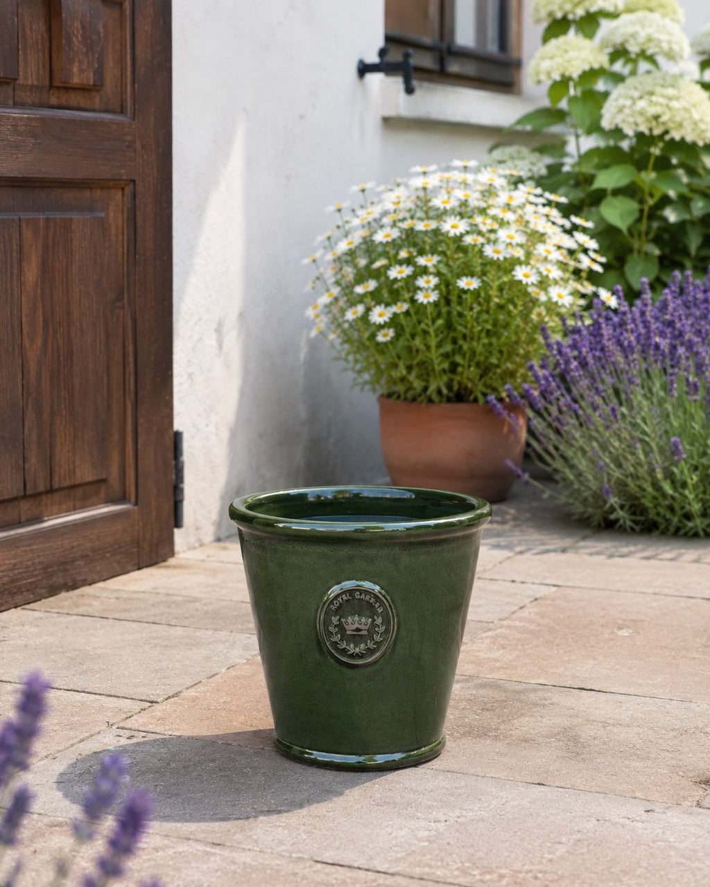 Der glasierte Pflanzkübel „Garden Green“ (Höhe 19–38 cm) steht auf einer Steinterrasse neben einer braunen Tür, umgeben von Gänseblümchen, blühendem Lavendel und Hortensien neben einer weißen Wand.