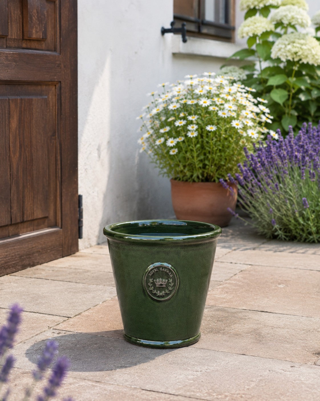 Der glasierte Pflanzkübel „Garden Green“ (Höhe 19–38 cm) steht auf einer Steinterrasse neben einer braunen Tür, umgeben von Gänseblümchen, blühendem Lavendel und Hortensien neben einer weißen Wand.