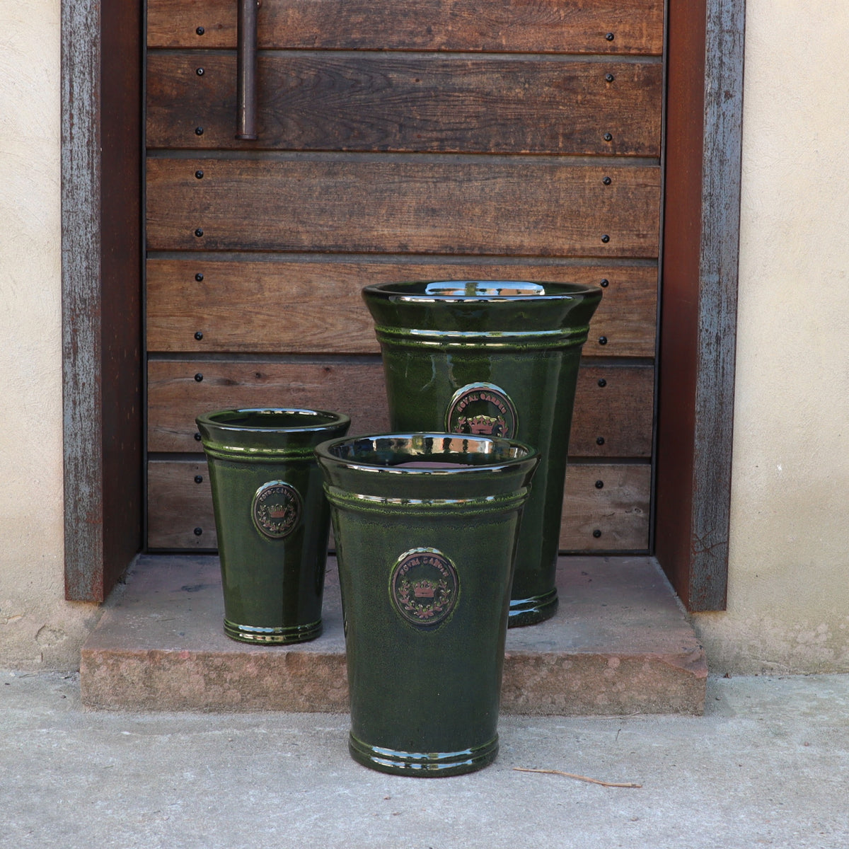 Drei "Royal Garden Green"-Vasen (Höhe 33-51 cm) stehen in unterschiedlichen Größen auf einer steinernen Türschwelle vor einer rustikalen Holztür mit horizontalen Bohlen und einem Metallgriff.