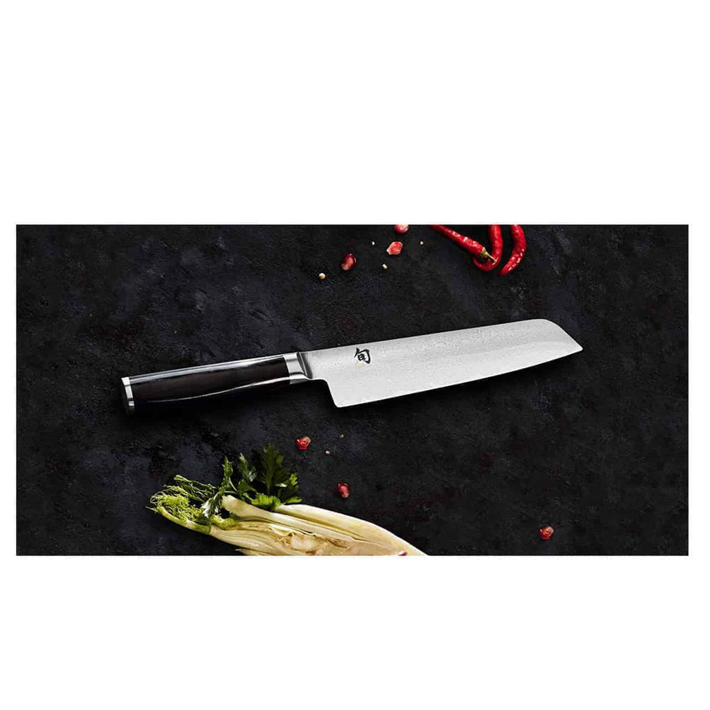 Ein hochwertiges Allzweckmesser, genauer gesagt das Kai Shun Premier Tim Mälzer Minamo TMM-0701 mit 15 cm ultrascharfer Klinge und dunklem Griff, liegt auf einer schwarzen Oberfläche. Frische Zutaten wie rote Chilischoten, Korianderblätter und ein Stück Pak Choy umgeben das Messer und suggerieren eine Kochvorbereitungsumgebung. In der Szene befindet sich auch ein Schneidebrett aus altem Fassholz (Eiche) mit den Maßen 25x15 cm2.
