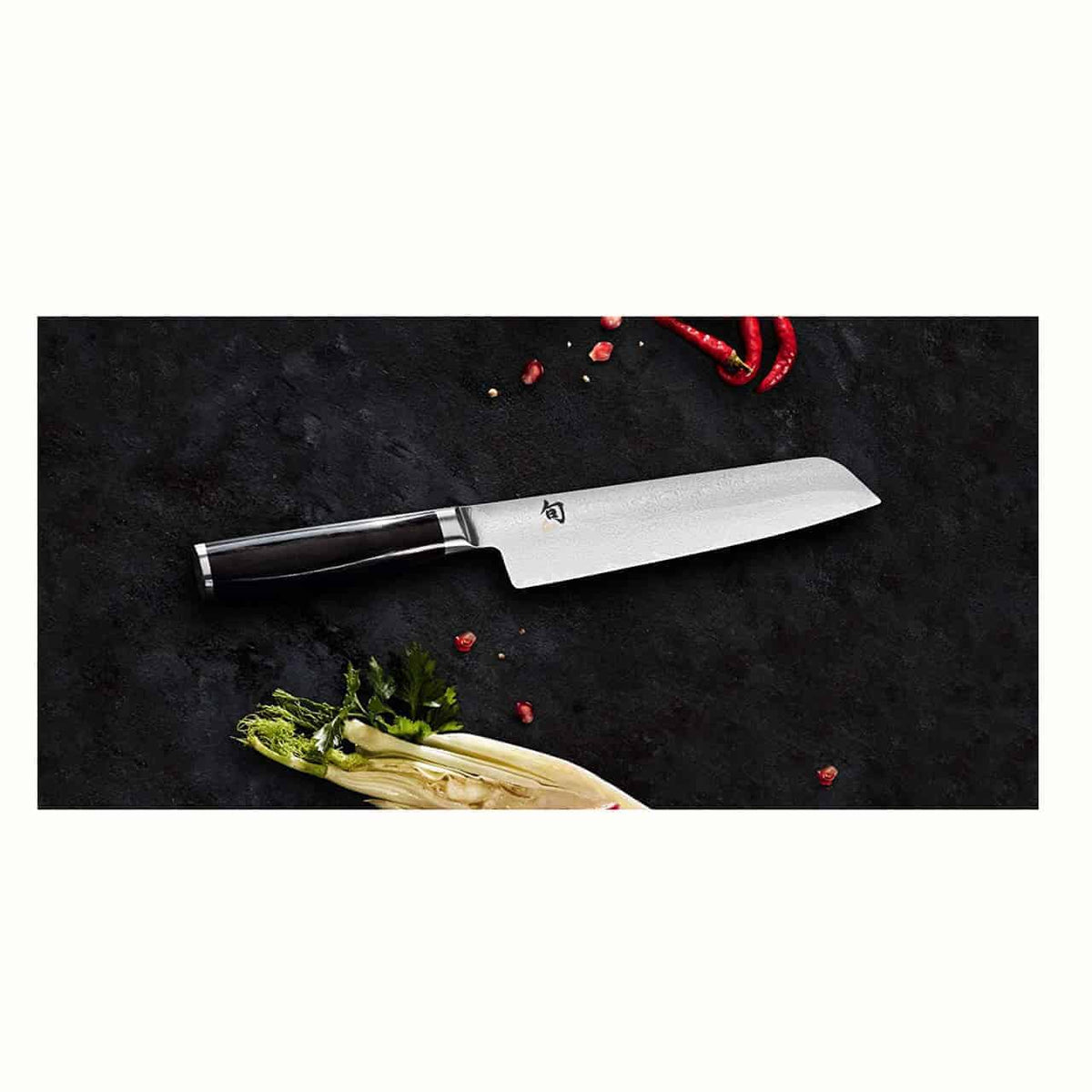 Ein Kai Shun Premier Minamo TMM-0700 Kochmesser mit schwarzem Griff und Damaszenerstahlklinge liegt auf einer dunklen Oberfläche. Um das Messer herum befinden sich rote Chilischoten, grüne Kräuter und ein teilweise sichtbares Blattgemüse. In der Nähe ruht ein Officemesser mit einer 9 cm langen Damaszenerstahlklinge auf einem rustikalen Küchenbrett Eiche mit den Maßen 30x22 cm.