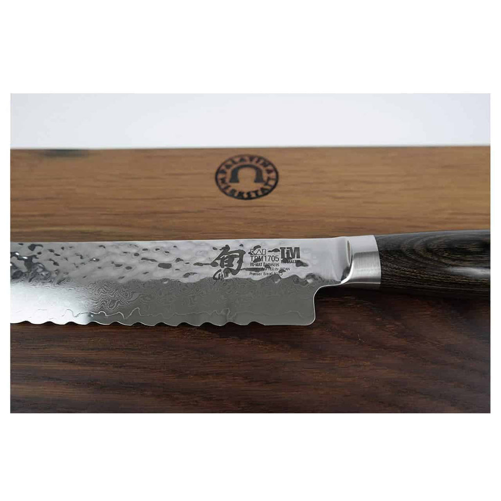 Eine Nahaufnahme eines Wellenschliffmessers mit aufwendig gemusterter Damaststahlklinge und Holzgriff aus dem KAI Shun Premier Tim Mälzer Geschenkset | TDM-1705. Das Messer ruht auf einem robusten Fassholzbrett mit den Maßen 30x18 cm. Auf der Klinge ist ein Logo mit den japanischen Schriftzeichen „TM 1705“ und „KAI Shun Premier“ zu sehen.