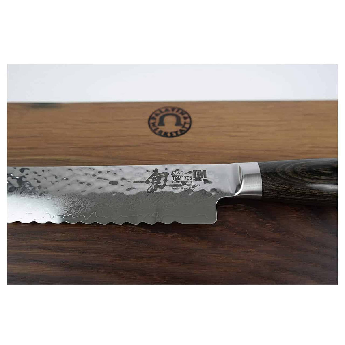 Eine Nahaufnahme eines Wellenschliffmessers mit aufwendig gemusterter Damaststahlklinge und Holzgriff aus dem KAI Shun Premier Tim Mälzer Geschenkset | TDM-1705. Das Messer ruht auf einem robusten Fassholzbrett mit den Maßen 30x18 cm. Auf der Klinge ist ein Logo mit den japanischen Schriftzeichen „TM 1705“ und „KAI Shun Premier“ zu sehen.