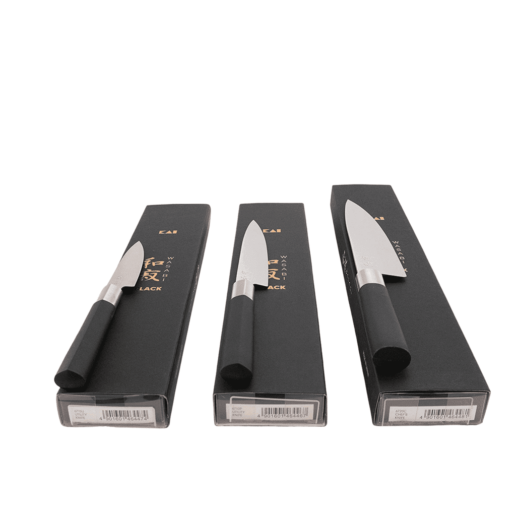 Eine Abbildung des Kai Wasabi Black Kochmesser-Sets zeigt drei Küchenmesser mit schwarzen Griffen, die jeweils elegant auf einer schwarzen Box angeordnet sind. Das Set enthält rechts ein 20 cm langes Kochmesser, in der Mitte ein 15 cm langes Allzweckmesser und links ein 10 cm langes Officemesser. Die Boxen sind mit Barcodes und etwas Text versehen. Zusätzlich ist ein 25x15 cm großes Schneidebrett aus Fassholz enthalten.