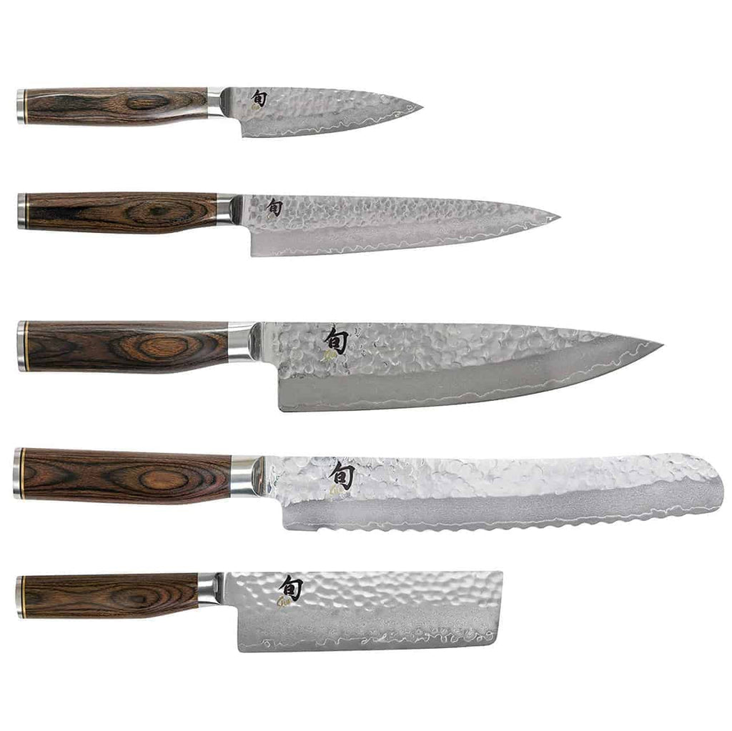 Ein Set aus fünf KAI Shun Tim Mälzer Damastmesser-Küchenmessern, untergebracht in einem Holzblock aus einem alten Weinfass, wird horizontal ausgestellt. Jedes Messer hat einen Holzgriff und eine gehämmerte Stahlklinge. Das Set umfasst ein kleines Schälmesser, ein Allzweckmesser, ein Kochmesser, ein Brotmesser und ein quadratisches Gemüsemesser.