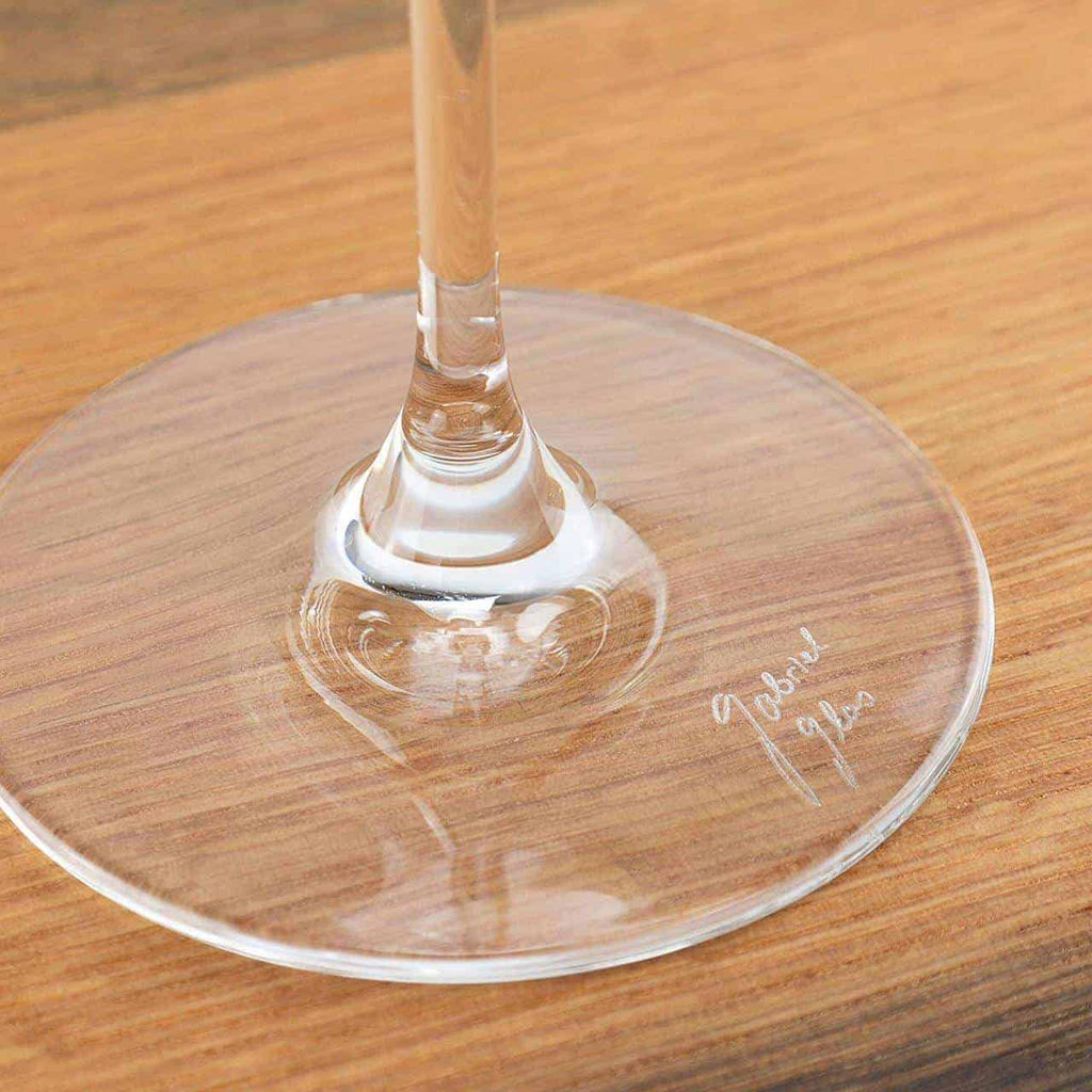 Eine Nahaufnahme zeigt den Boden eines klaren Weinglases, das auf einer Holzoberfläche ruht. In den Glasboden ist eine Autogrammangabe mit Jahreszahl eingraviert. Das Foto hebt das Design des Glases und die detaillierte Inschrift hervor.