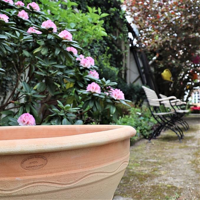 Nahaufnahme eines großen, leeren winterfesten Pflanzkübels, Blumenkübel 70 cm, Artemisia in einem Garten mit rosa blühenden Blumen und grünem Laub im Hintergrund. Entlang eines Weges stehen Bänke und man sieht Bäume mit roten Blättern. Die Szene wirkt friedlich und gepflegt.