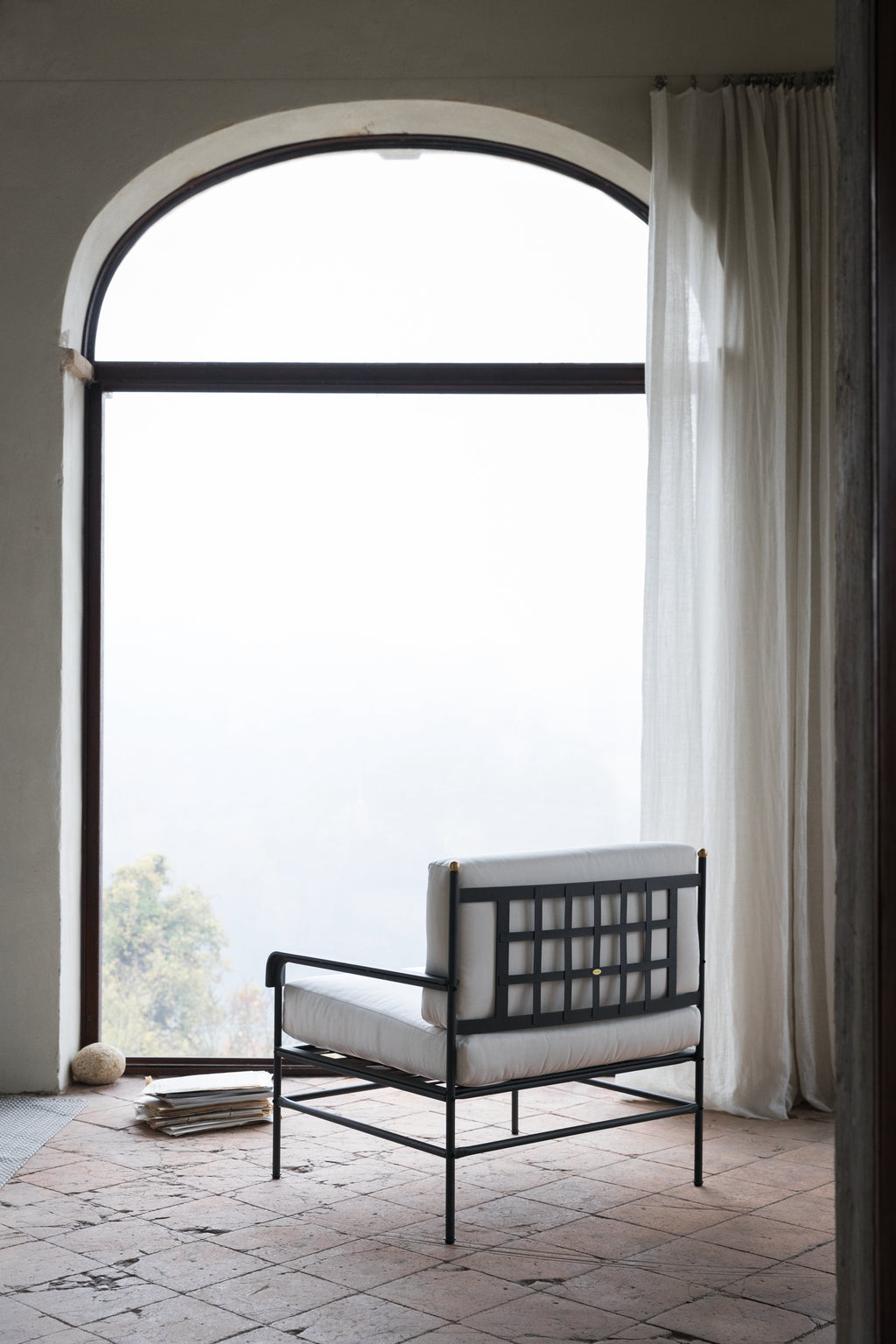 Ein minimalistischer Raum mit einem bogenförmigen Fenster, halbtransparenten weißen Vorhängen und einem modernen Sessel Toscana aus Eisen in Graphitgrau mit weißen Kissen. Der Raum hat einen rustikalen Fliesenboden und einen Blick auf eine neblige Landschaft draußen. Ein kleiner Stapel Bücher und ein Wollknäuel liegen auf dem Boden.