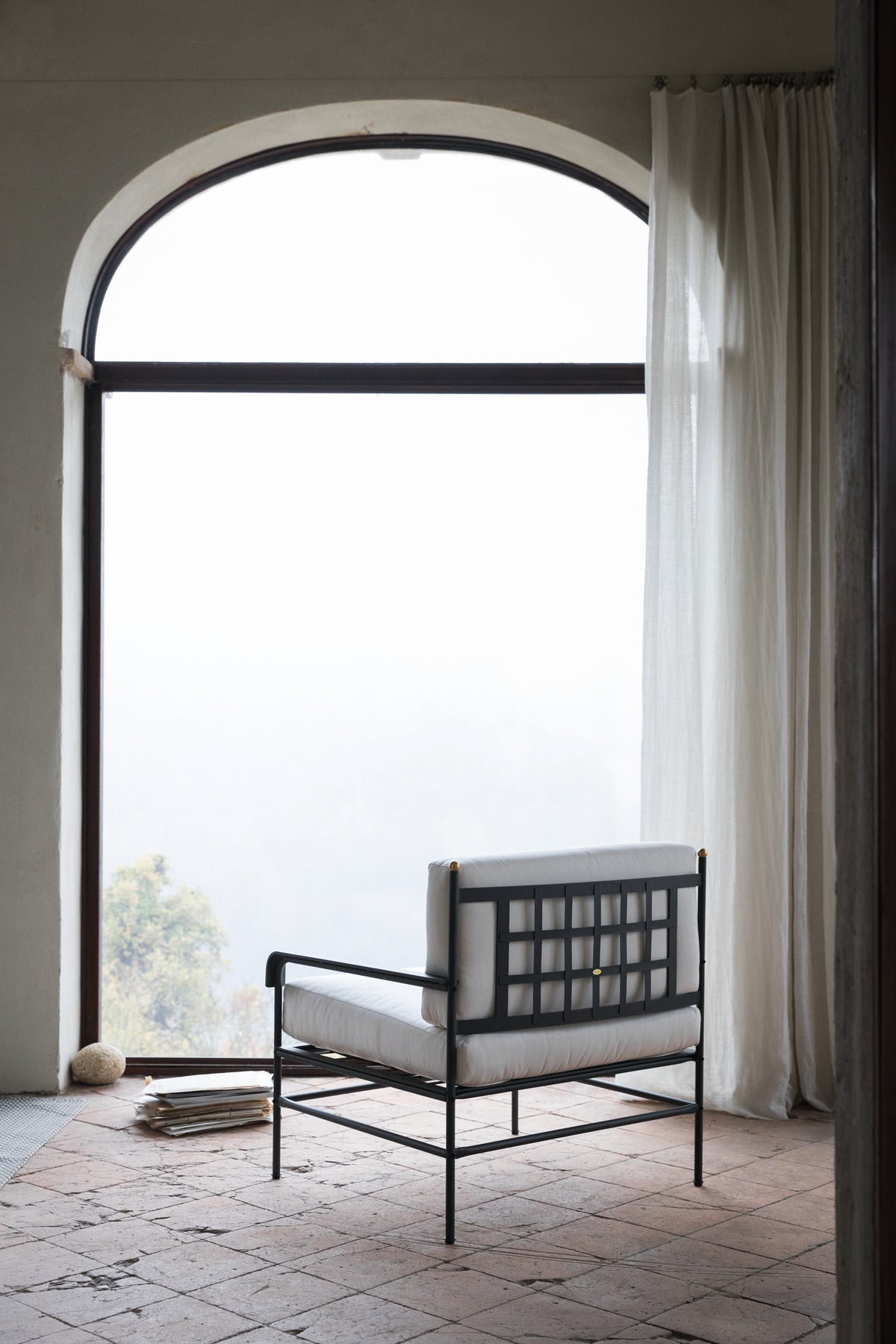 Ein minimalistischer Raum mit einem bogenförmigen Fenster, halbtransparenten weißen Vorhängen und einem modernen Sessel Toscana aus Eisen in Graphitgrau mit weißen Kissen. Der Raum hat einen rustikalen Fliesenboden und einen Blick auf eine neblige Landschaft draußen. Ein kleiner Stapel Bücher und ein Wollknäuel liegen auf dem Boden.