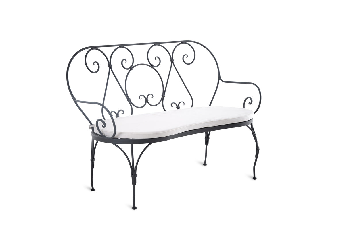 Eine dekorative Sitzbank Aurora aus schwarzem Schmiedeeisen mit weißem Kissen. Das Design umfasst elegante Schnörkel an der Rückenlehne und den Armlehnen, wodurch sie sich perfekt für den Einsatz im Freien eignet und gleichzeitig Funktionalität und Ästhetik vereint.