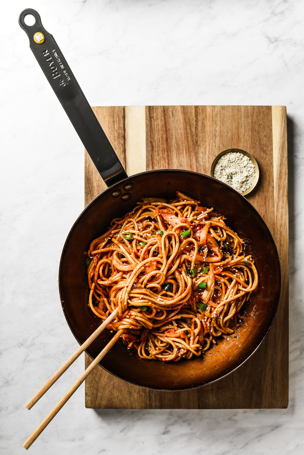 Eine Pfanne mit Nudeln in roter Soße mit Kräutern steht auf einem Holzbrett, auf dem der De Buyer Wok Mineral B, 28 cm Dm steht. Im Wok liegen Essstäbchen und eine kleine Schale mit Gewürzen, perfekt für die authentische asiatische Küche.