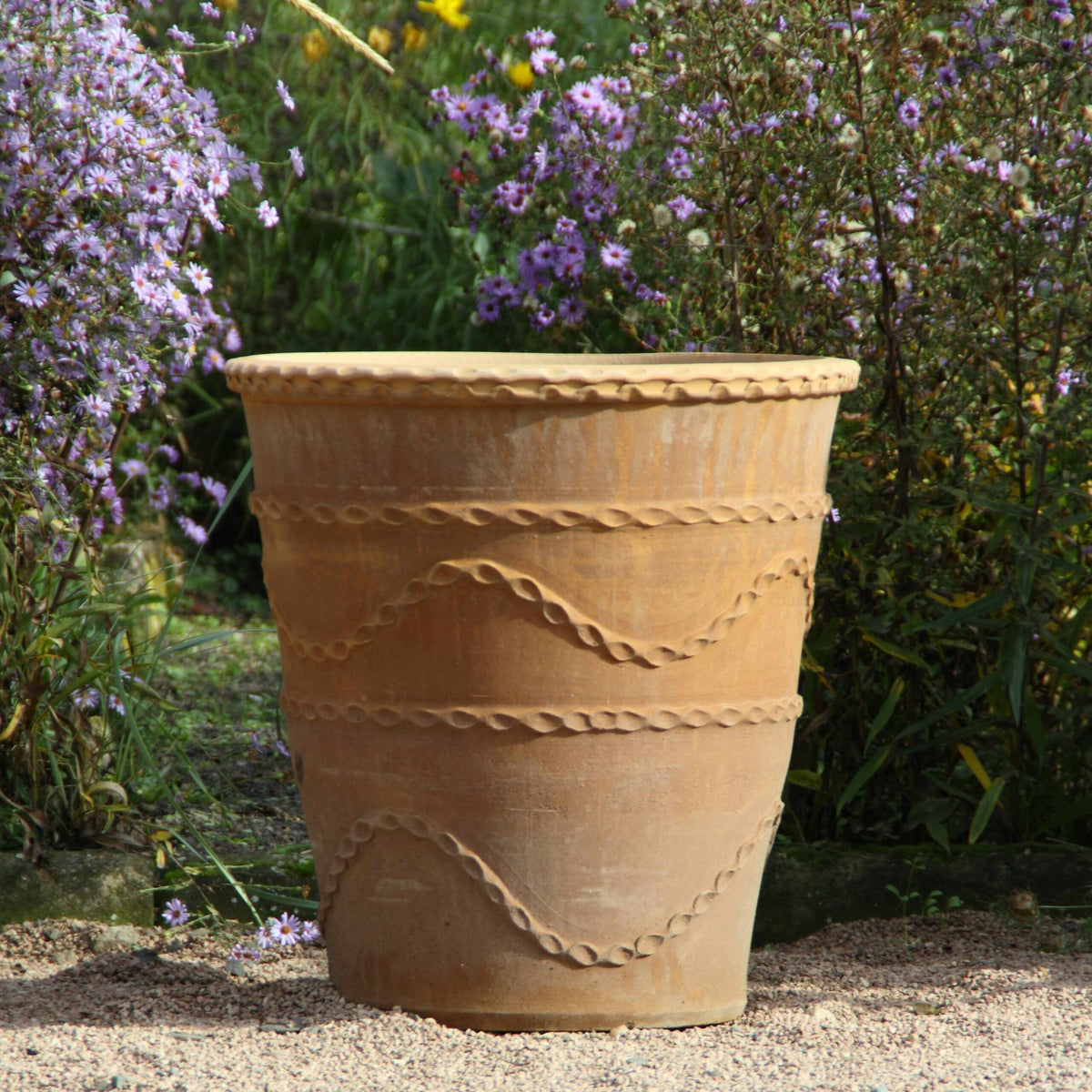Ein Terrakotta-Pflanzkübel „Acanthus“ mit dekorativem Wellenmuster steht auf einem Kiesweg. Im Hintergrund zieren üppiges Grün und blühende lila Acanthus-Blüten eine lebendige Gartenszene.