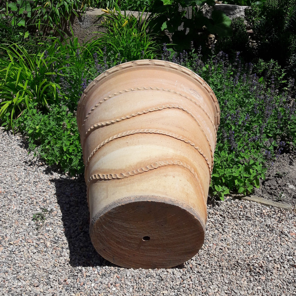 Ein großer, leerer Terrakotta-Pflanzkübel „Acanthus“ (H=50 cm) liegt auf der Seite in einem kiesigen Gartenbereich. Der Topf weist dekorative Rillen und ein Wellenmuster auf seiner Oberfläche auf und ist von grünem Laub und Acanthus-Pflanzen umgeben.