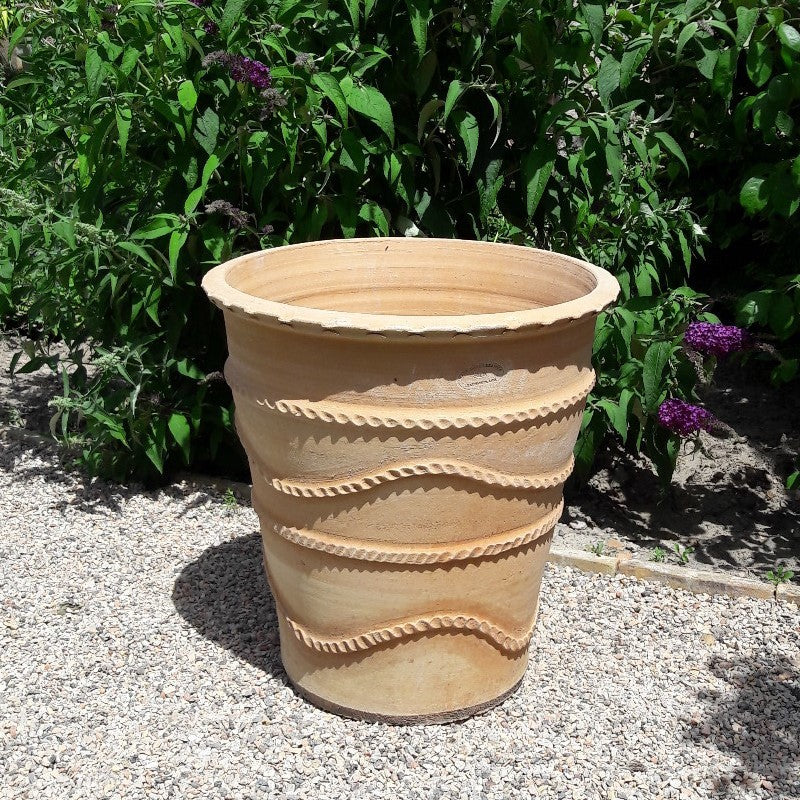 Ein Pflanzkübel „Acanthus“ aus Terrakotta mit wellenförmigem Rillenmuster steht auf einer Kiesfläche. Im Hintergrund ist dichtes grünes Laub zu sehen, einige Acanthusblüten sind zu erkennen. Der Pflanzkübel ist leer und steht im Sonnenlicht.