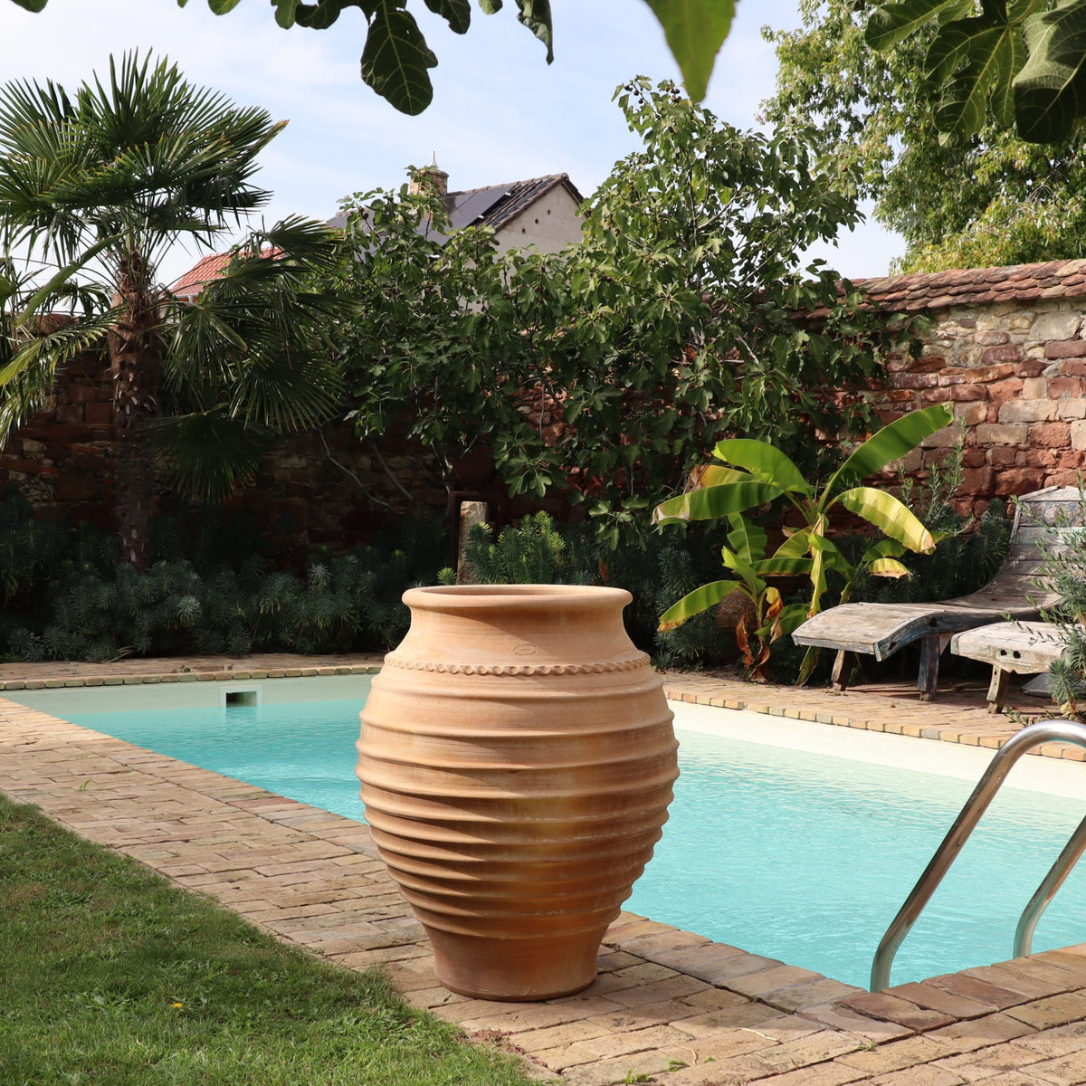 Die frostfeste Amphore "Agave", H=70cm, steht an einem Pool mit Steinfliesen, üppigem Grün, Bananenstauden und Liegestühlen in einem sonnigen, von Mauern umgebenen Garten.