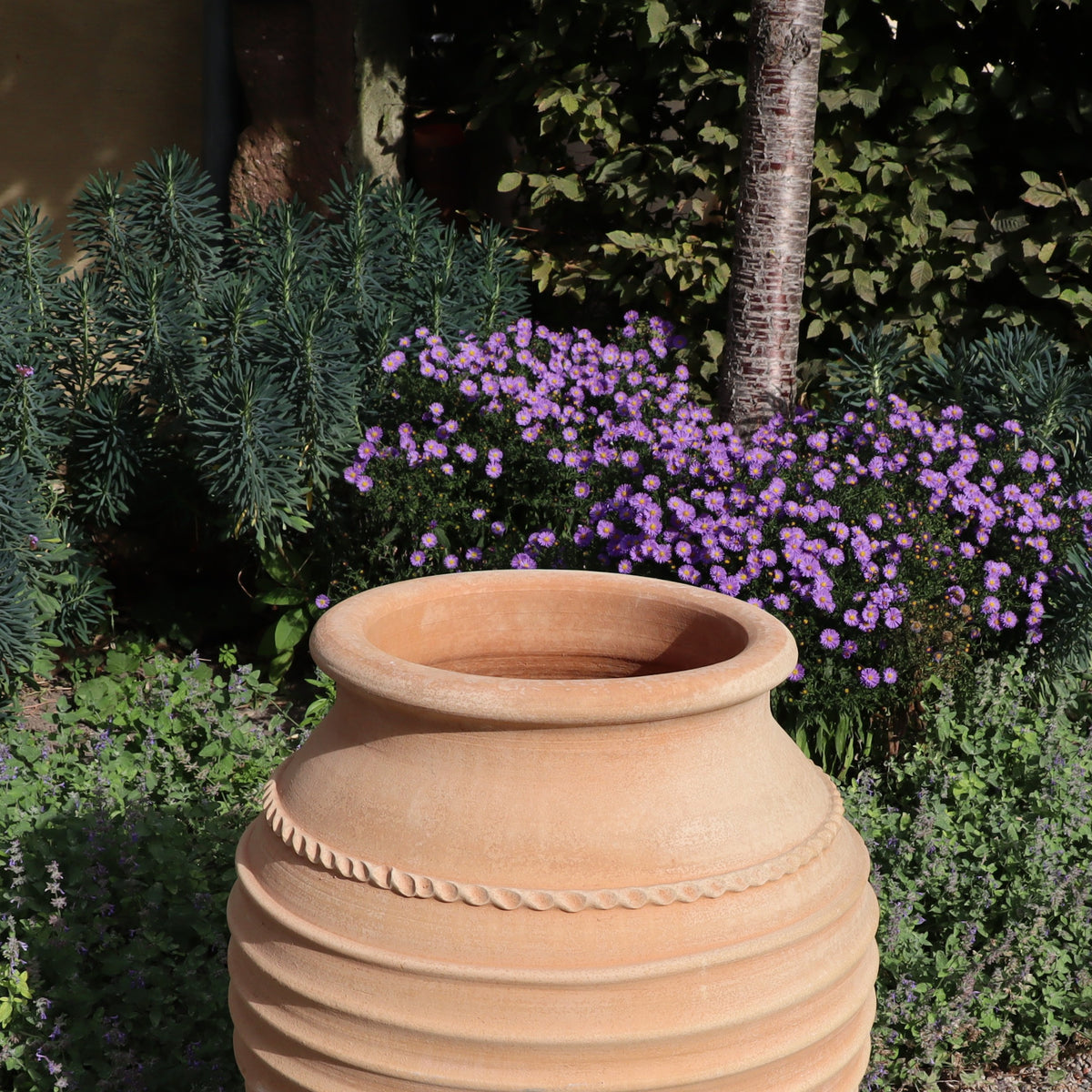 Eine frostfeste Amphore "Agave", H=80cm, steht im Garten zwischen grünem Laub und violetten Blüten, mit einem Baumstamm und belaubten Büschen im Hintergrund.
