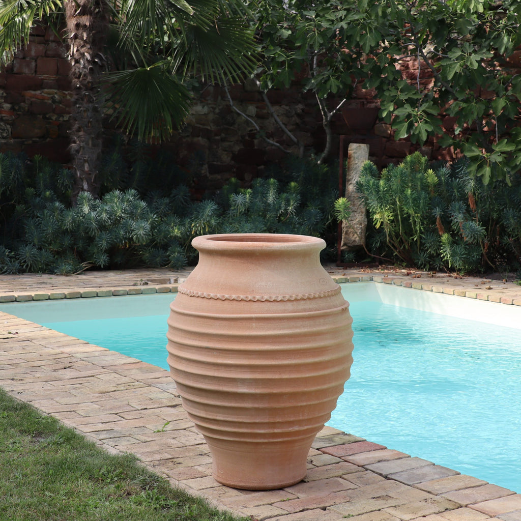 Eine frostfeste Amphore "Agave", H=80cm, steht auf einer Backsteinterrasse an einem klaren blauen Pool, umgeben von üppigen grünen Pflanzen und Bäumen.