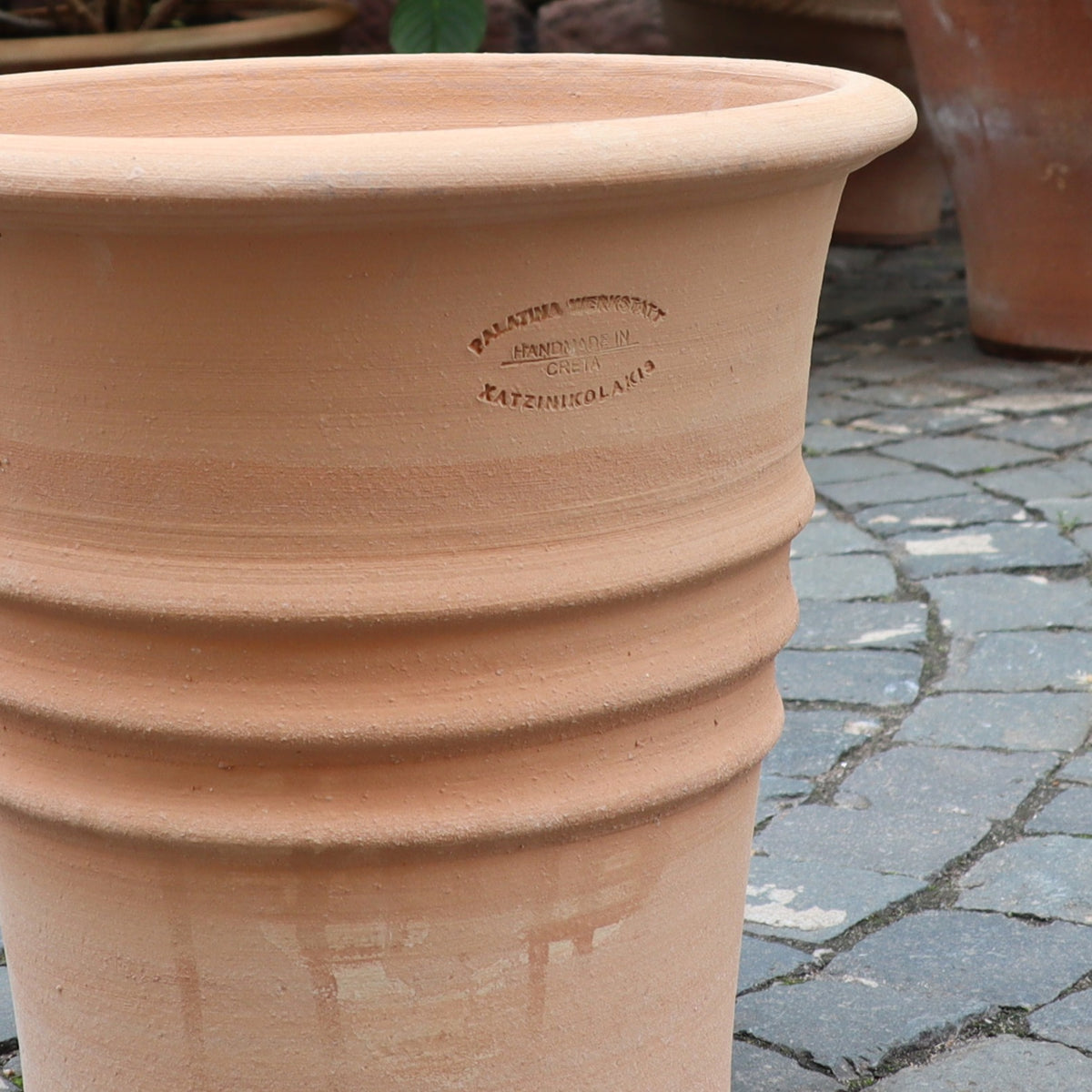 Der Blumentopf "Aster" H=35 cm, ein großer Terrakotta-Pflanztopf mit ausgestelltem Rand, Rillen und Seitenstempel, steht leer auf einem Steinpflaster mit weiteren Pflanzgefäßen und Grünpflanzen im Hintergrund.