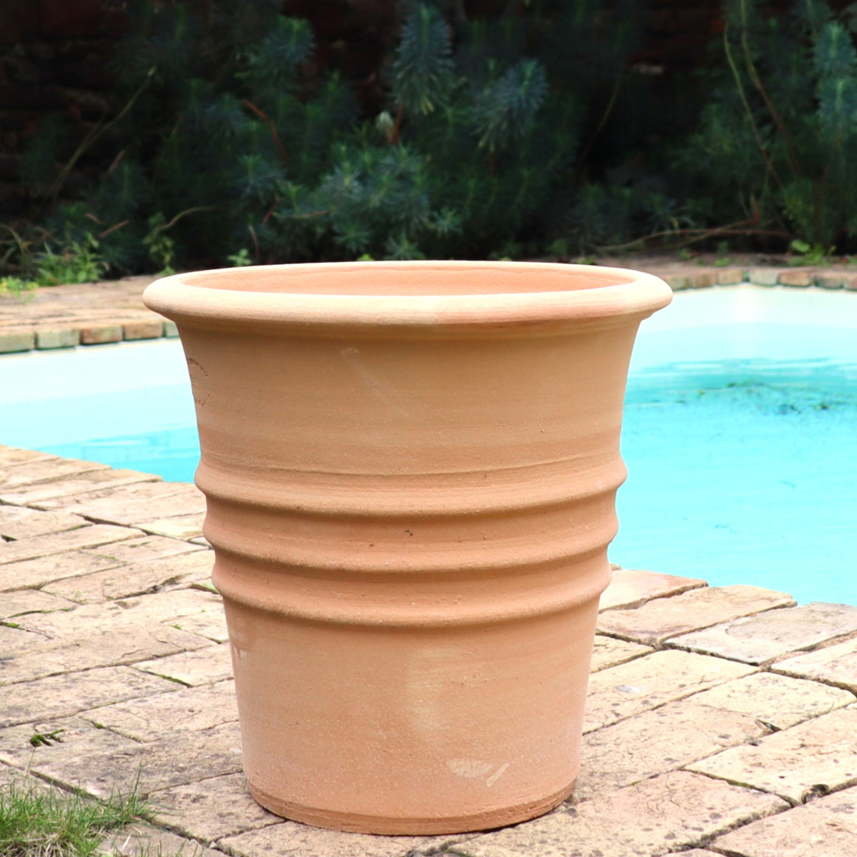 Der Blumentopf "Aster" H=35 cm, ein großer Terrakotta-Pflanzkübel mit geriffelten Rändern und einem Entwässerungsloch, steht auf einer gemauerten Terrasse neben einem Schwimmbad, mit üppigem Grün im Hintergrund.