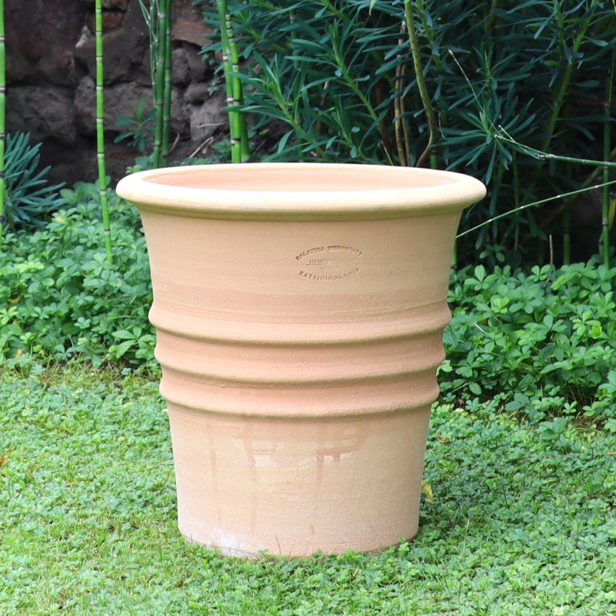 Ein großer, hellbrauner Blumentopf "Aster" (H=35 cm) mit horizontalen Rillen und Entwässerungsloch sitzt auf grünem Gras, umgeben von üppigen Pflanzen und Bambus im Hintergrund.
