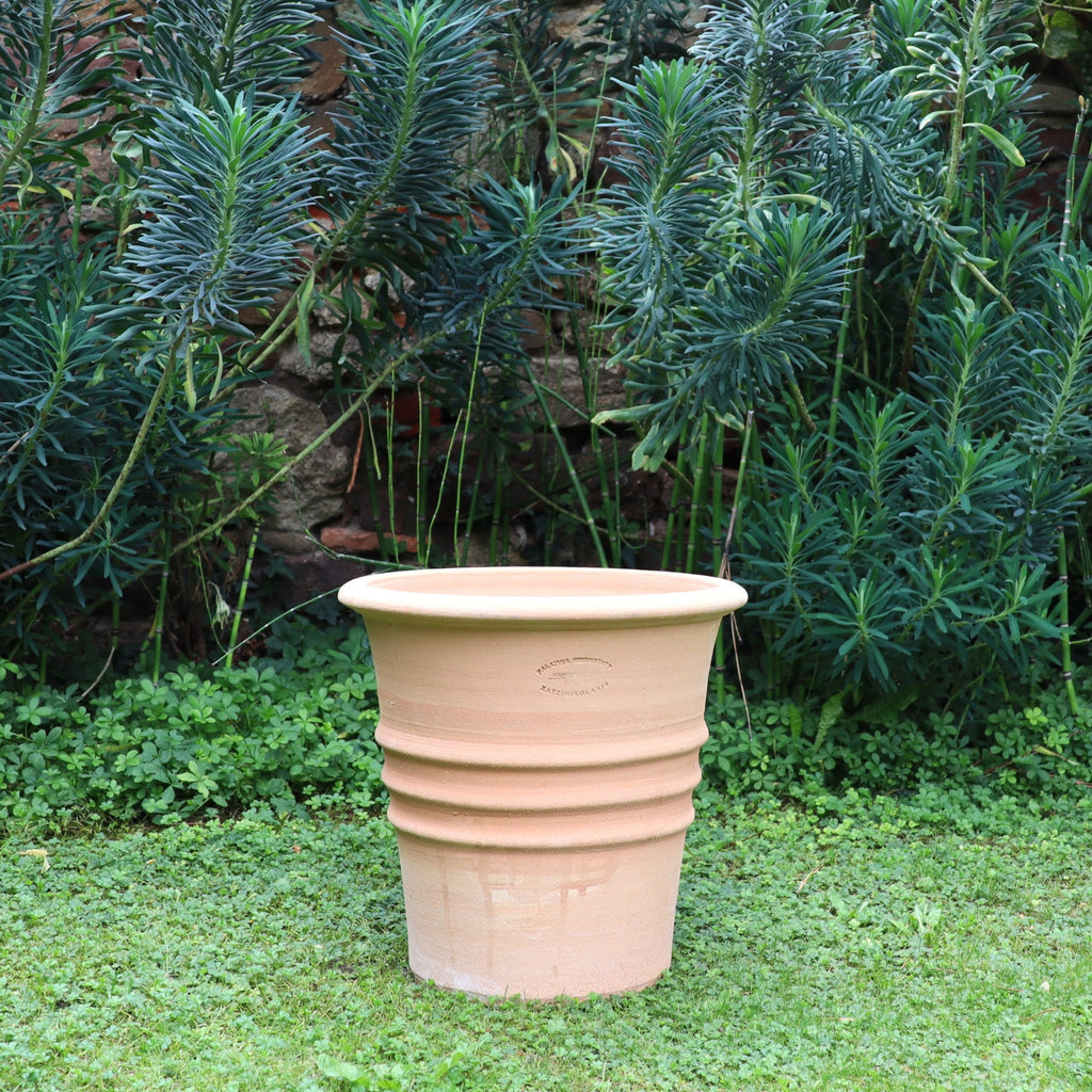 Der Blumentopf "Aster" H=35 cm aus Terrakotta mit Auslaufloch und gerillten Details steht auf grünem Rasen, umgeben von hohen Pflanzen und einer rustikalen Ziegelwand im Hintergrund.