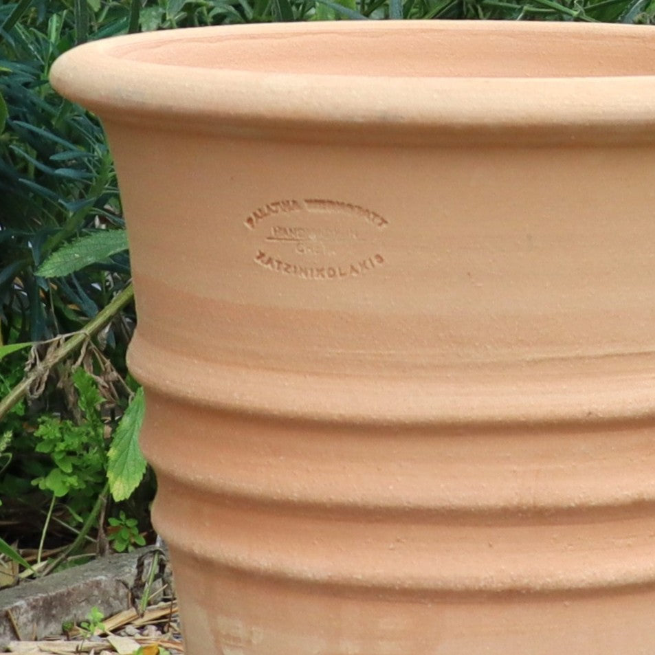 Nahaufnahme des großen, geriffelten Blumentopf "Aster" H=35 cm Terrakotta-Pflanzgefäßes mit Herstellerstempel, das im Freien zwischen Grünpflanzen und Blättern steht.