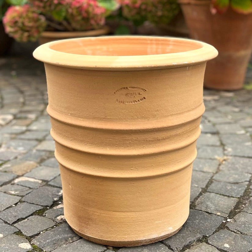 Ein großer Terrakotta-Blumentopf "Aster", H=40 cm, mit drei horizontalen Rillen steht auf einer Pflastersteinfläche. Andere Terrakottatöpfe und Grünpflanzen erscheinen unscharf im Hintergrund.