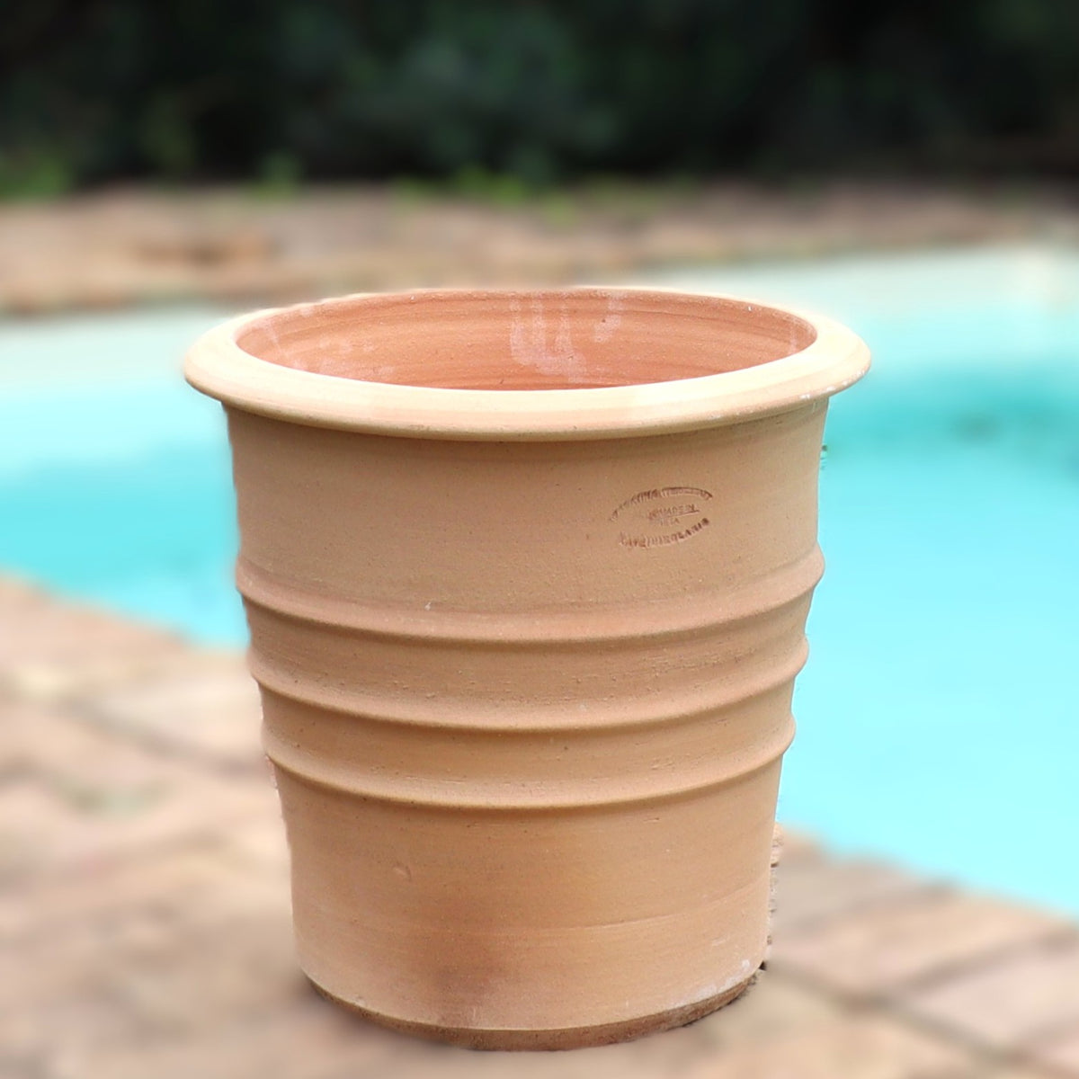 Ein großer, leerer Terrakotta-Blumentopf "Aster", H=40 cm, steht auf einer Ziegelfläche neben einem Swimmingpool mit türkisfarbenem Wasser und Grün im Hintergrund.
