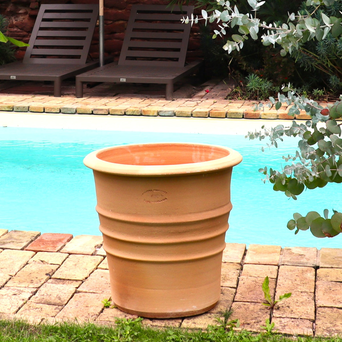 Ein großer, leerer Terracotta Blumentopf "Aster", H=40 cm, steht auf einer gemauerten Terrasse neben einem Swimmingpool, mit Liegestühlen und üppigem Grün im Hintergrund.