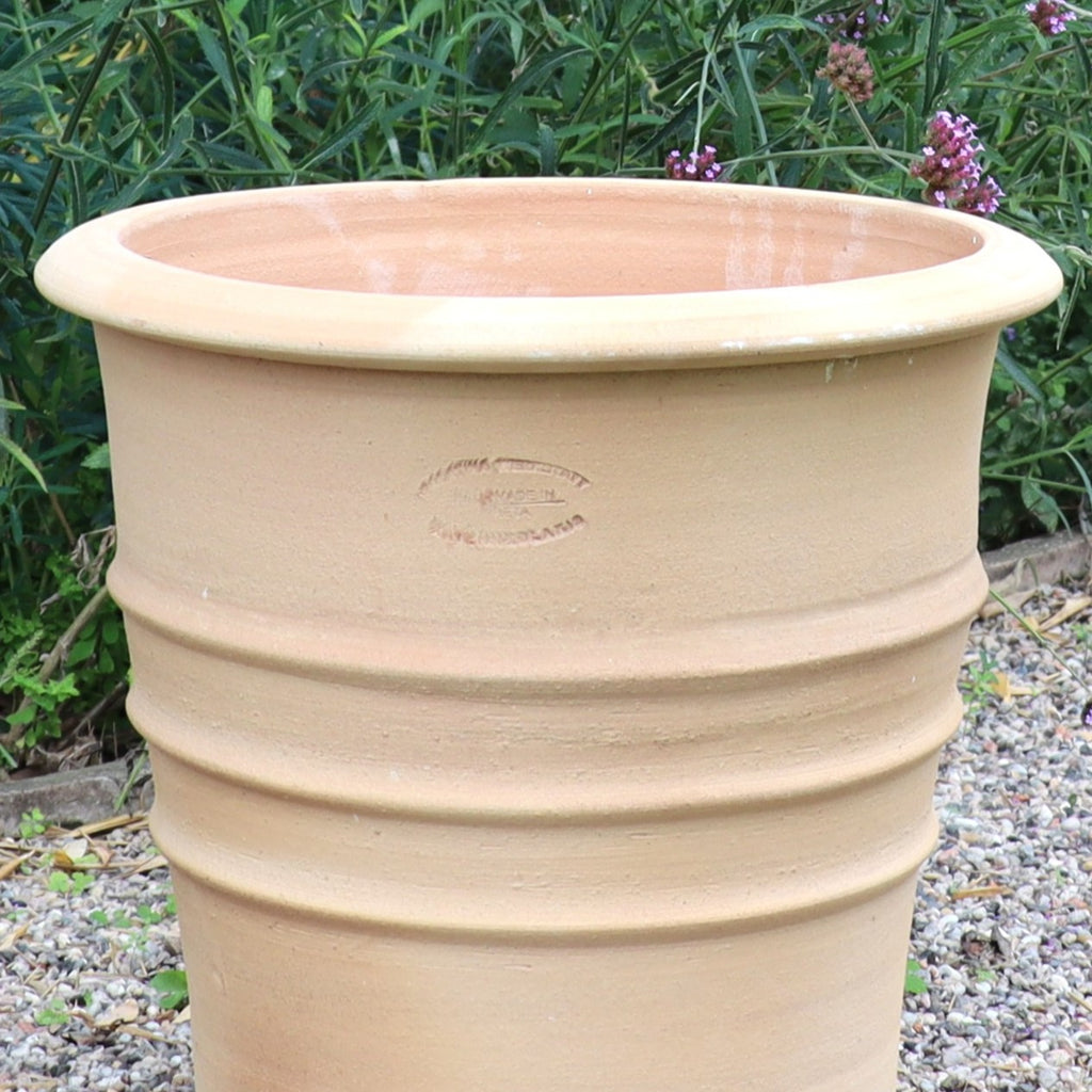 Der Terracotta Blumentopf "Aster", H=40 cm, mit feinen Rillen, steht unglasiert auf einem Kiesbett, umgeben von grünen Blattpflanzen und lila Blüten im Hintergrund.