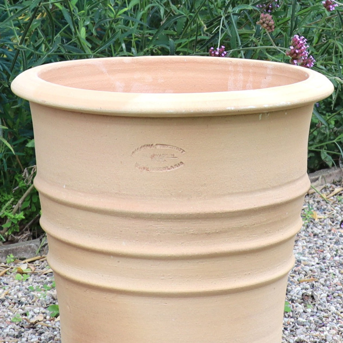 Der Terracotta Blumentopf "Aster", H=40 cm, mit feinen Rillen, steht unglasiert auf einem Kiesbett, umgeben von grünen Blattpflanzen und lila Blüten im Hintergrund.